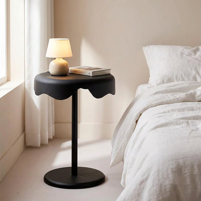 Black round side table used as bedside table next to bed with lamp and book in modern bedroom