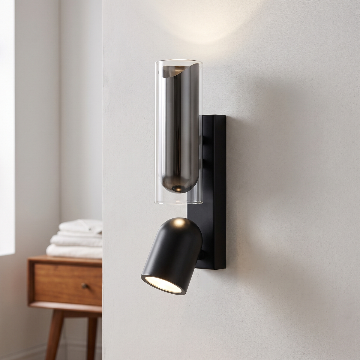 Matte black up and down spotlight wall light with glass tube up-light, lit beside a bed