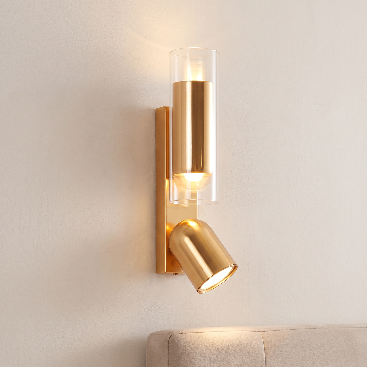 Brass up and down spotlight wall light with glass tube up-light, lit on a plaster wall beside a bed